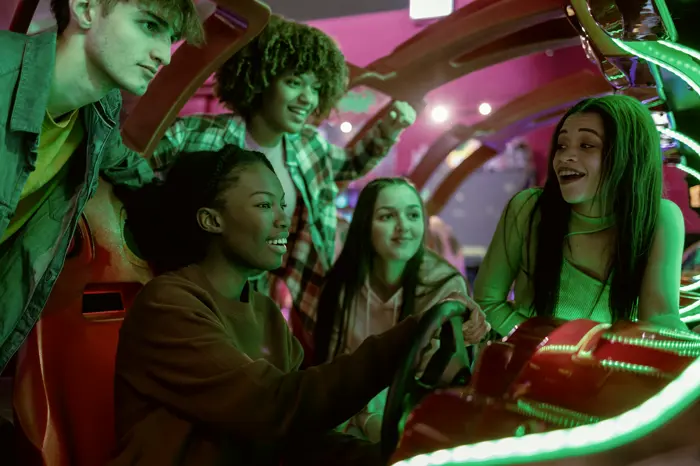 A group of friends crowded around a racing cabinet, one on the wheel, the rest coaching loudly.
