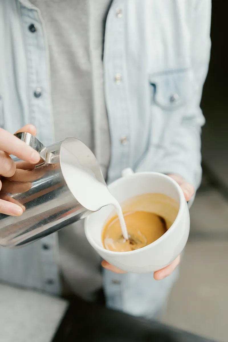 A barista pouring steamed milk from a steel jug into a white cup of espresso.