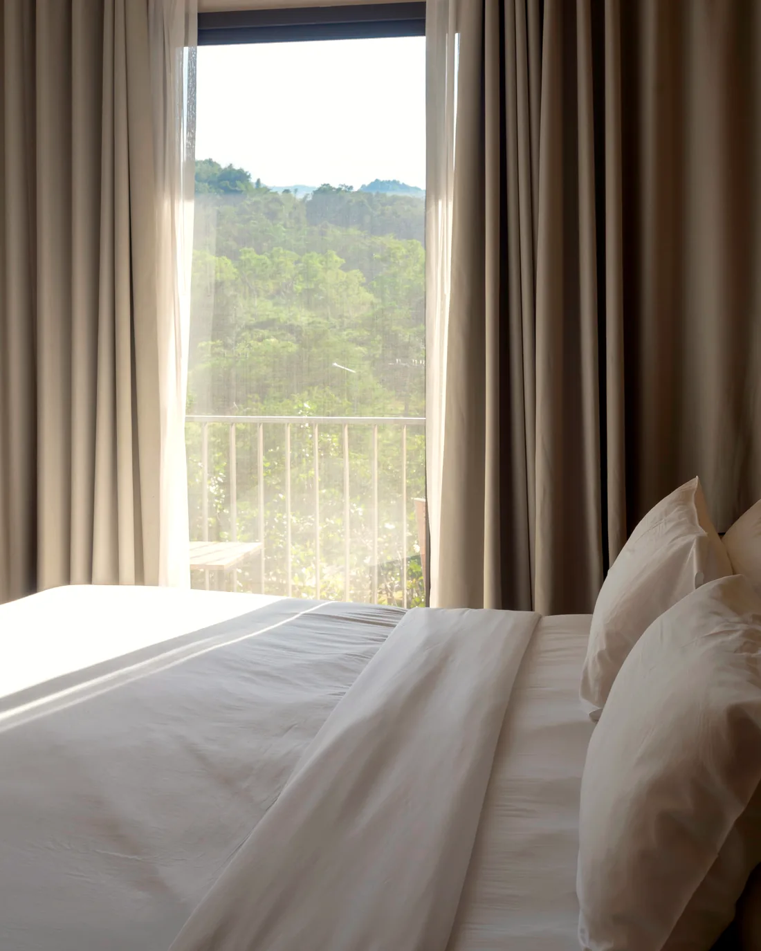 Soft light through half-drawn curtains onto white bedding — the quiet hour before the table is laid.