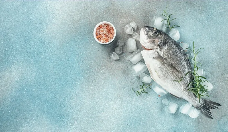 A whole dorade laid on pale blue stone among scattered ice, a small bowl of pink salt beside it.