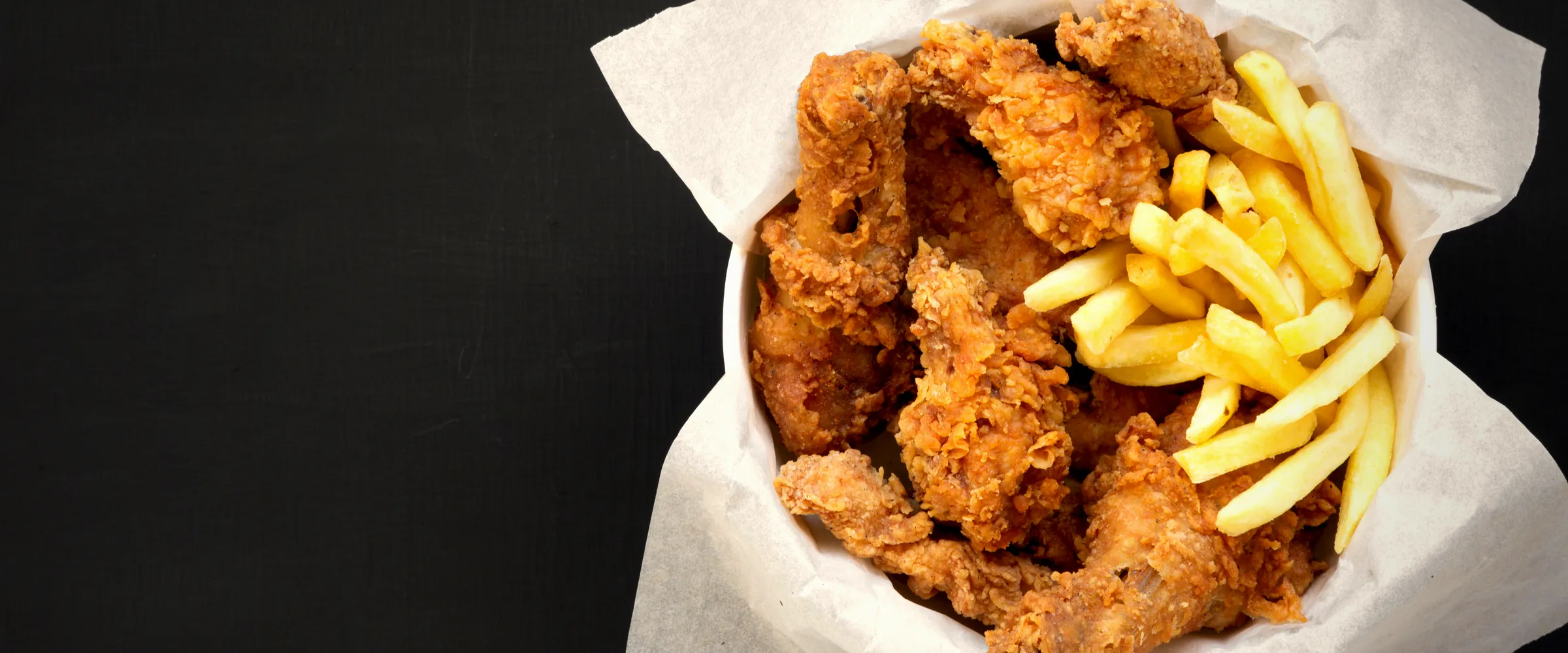 A paper-lined basket of fried chicken pieces and fries, shot from above on black.