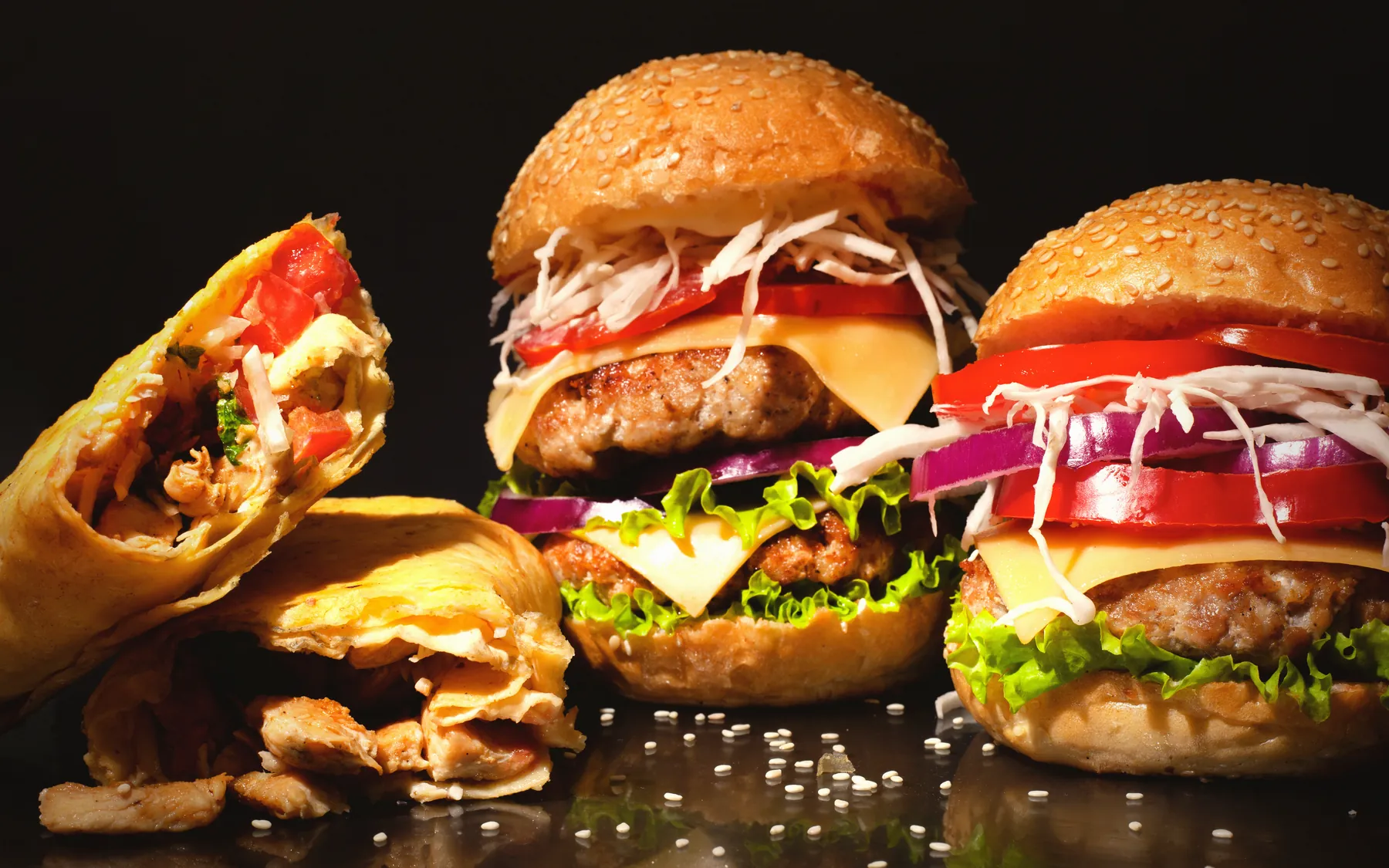 Several stacked burgers and a loaded wrap grouped on a dark reflective counter.