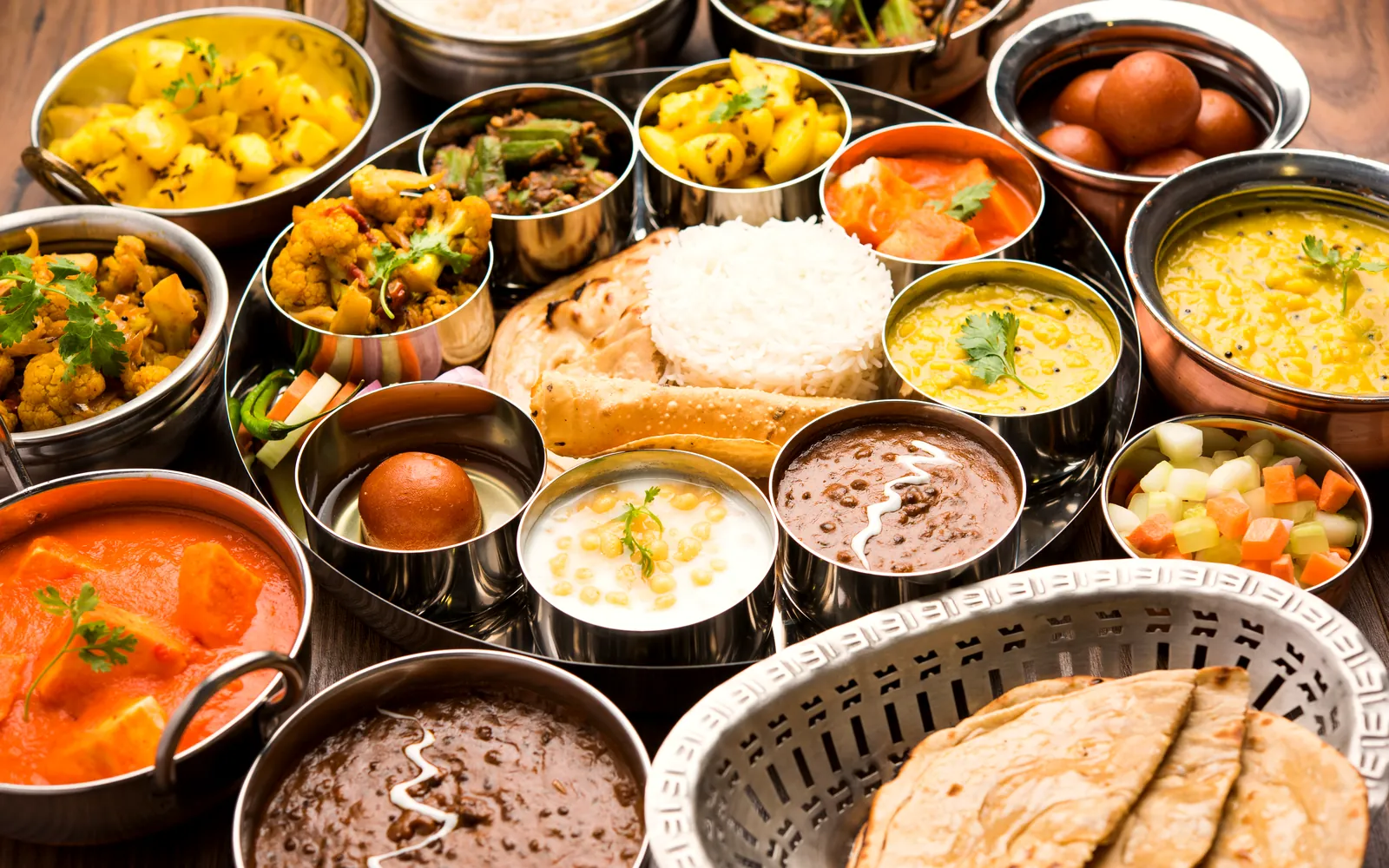 A table crowded with steel katoris: curries, dal, rice, breads and sweets making up a full thali.