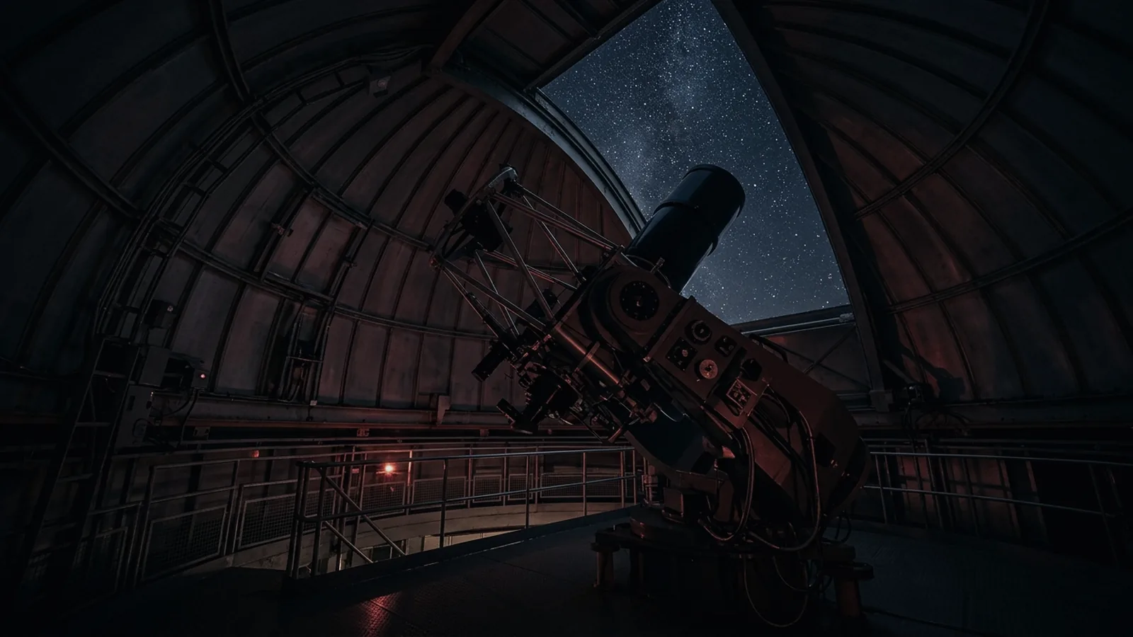Inside the observatory dome at night: a large reflecting telescope angled up through the open shutter slit, with a wedge of star field visible and the curved dome wall lit only by dim red lamps.