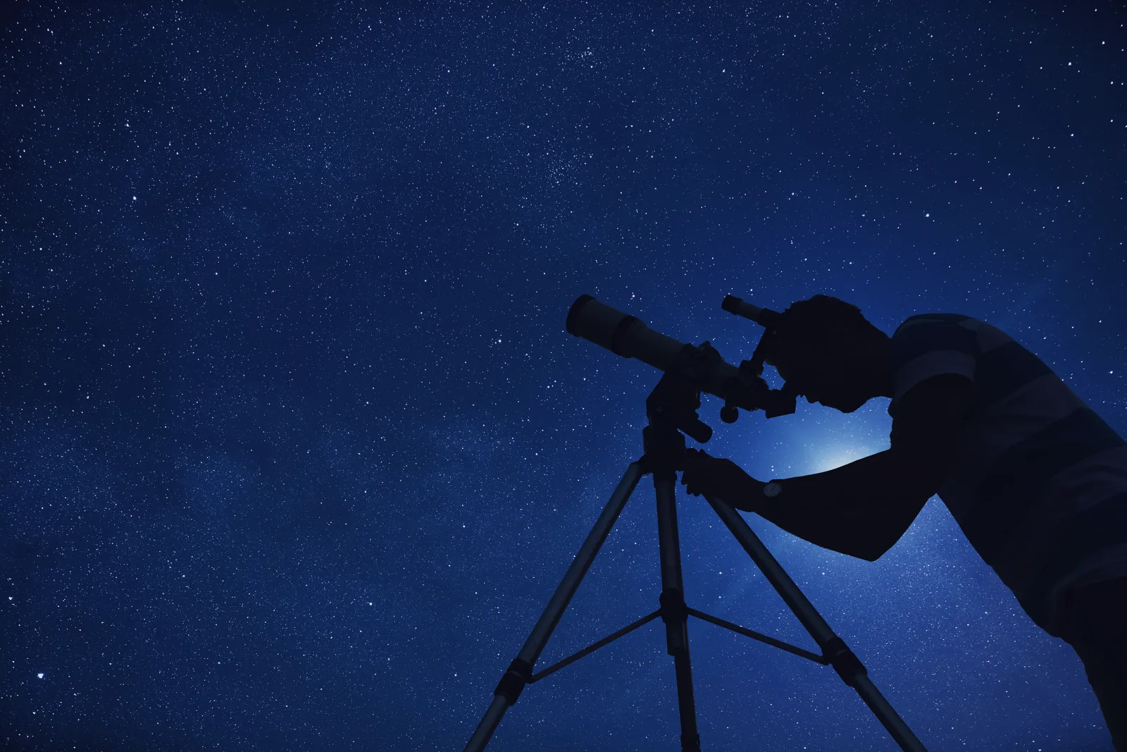 A person leaning to the eyepiece of a telescope on a tripod, silhouetted against a deep blue star-filled sky.