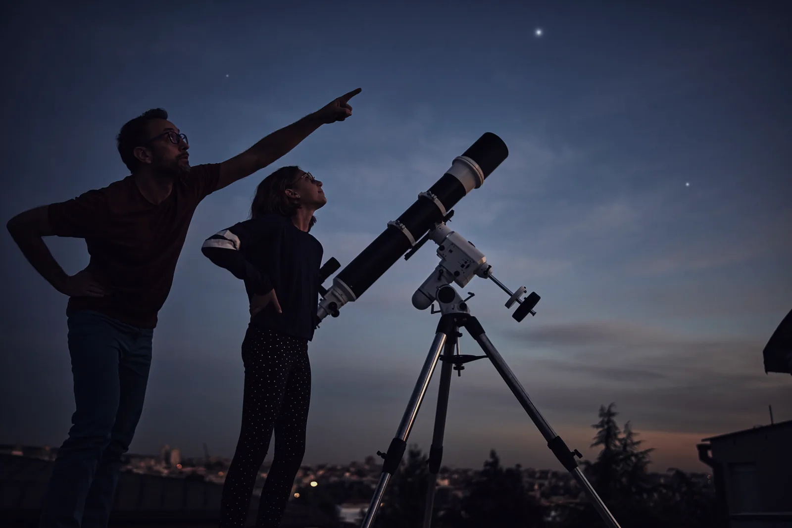 An adult and a child in silhouette beside a small telescope on a tripod, one of them pointing up at a deep blue twilight sky.