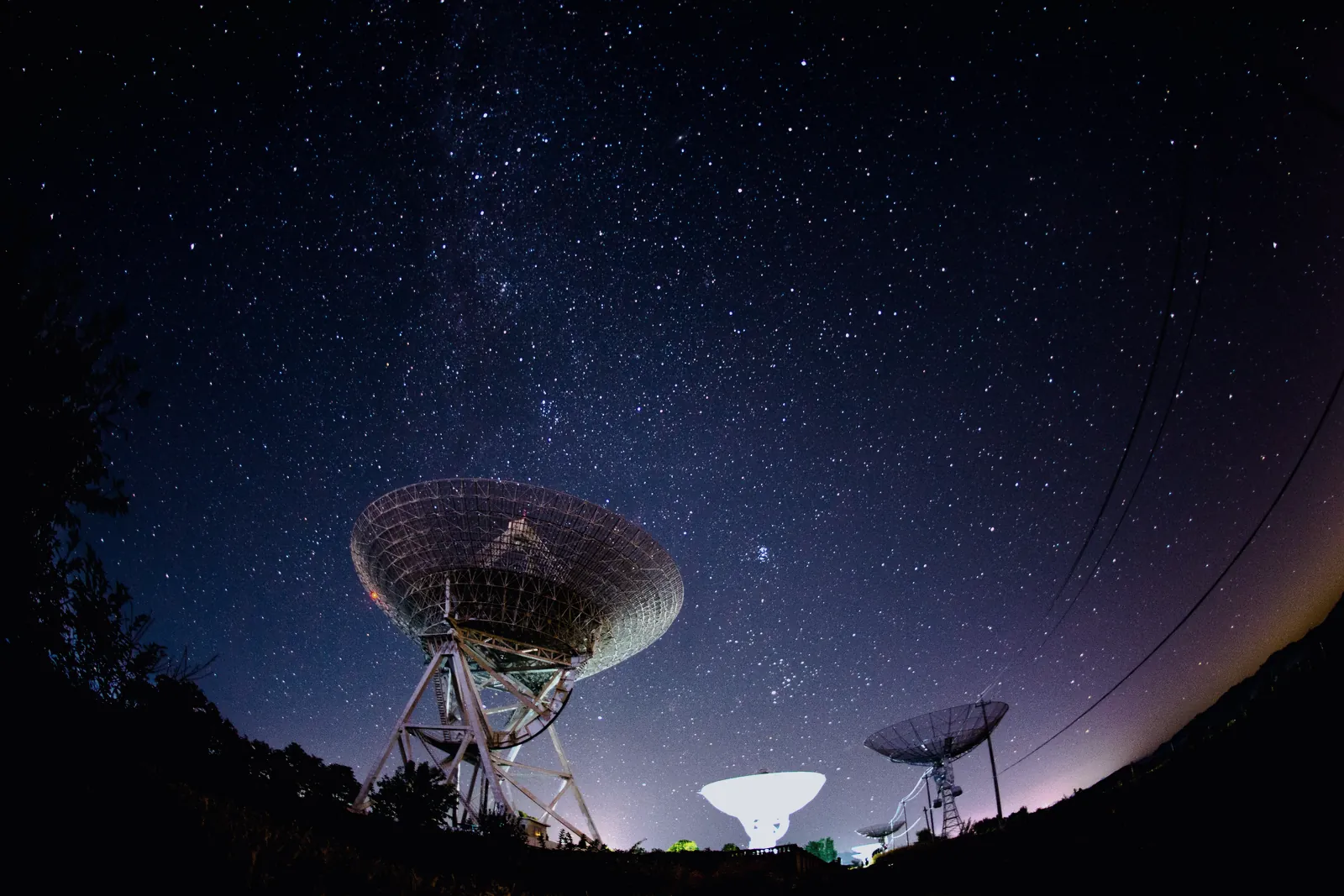 Radio telescope dishes silhouetted against a star-filled night sky, with the Milky Way overhead and a faint glow along the horizon.