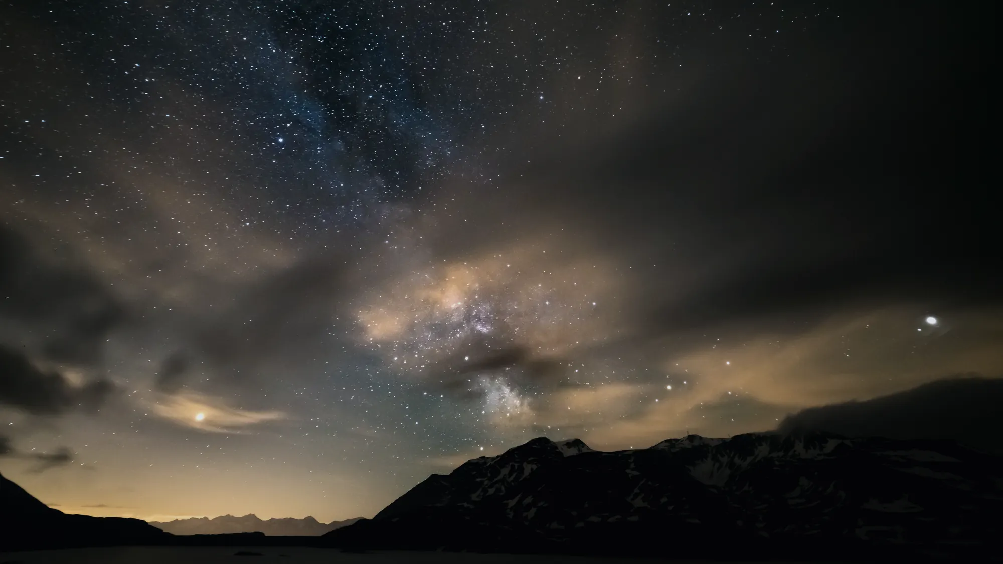 The Milky Way over a dark mountain ridge with low cloud below, and a faint orange glow of distant town lighting on the horizon.