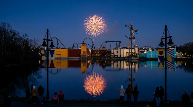 A single firework bursting over the darkened park skyline, reflected in the lake, watched from the far shore.