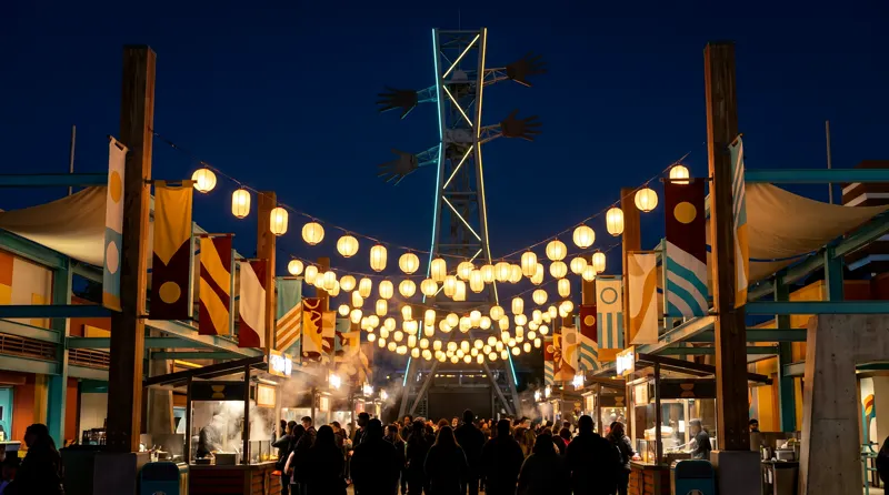 The Yard at night: lantern strings over the Night Market, the Hundredhand tower edge-lit in cyan above the crowd.
