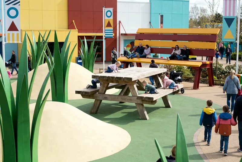Little Giants: children climbing an oversized picnic table beneath sculpted three-metre grass blades, parents on a giant bench behind.