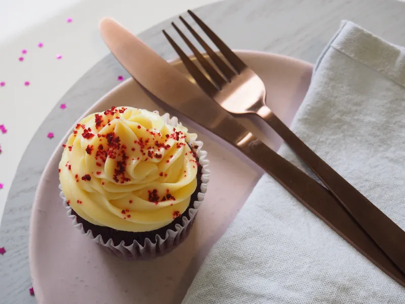 A cupcake with pale frosting on a pink plate beside copper cutlery, photographed from above.