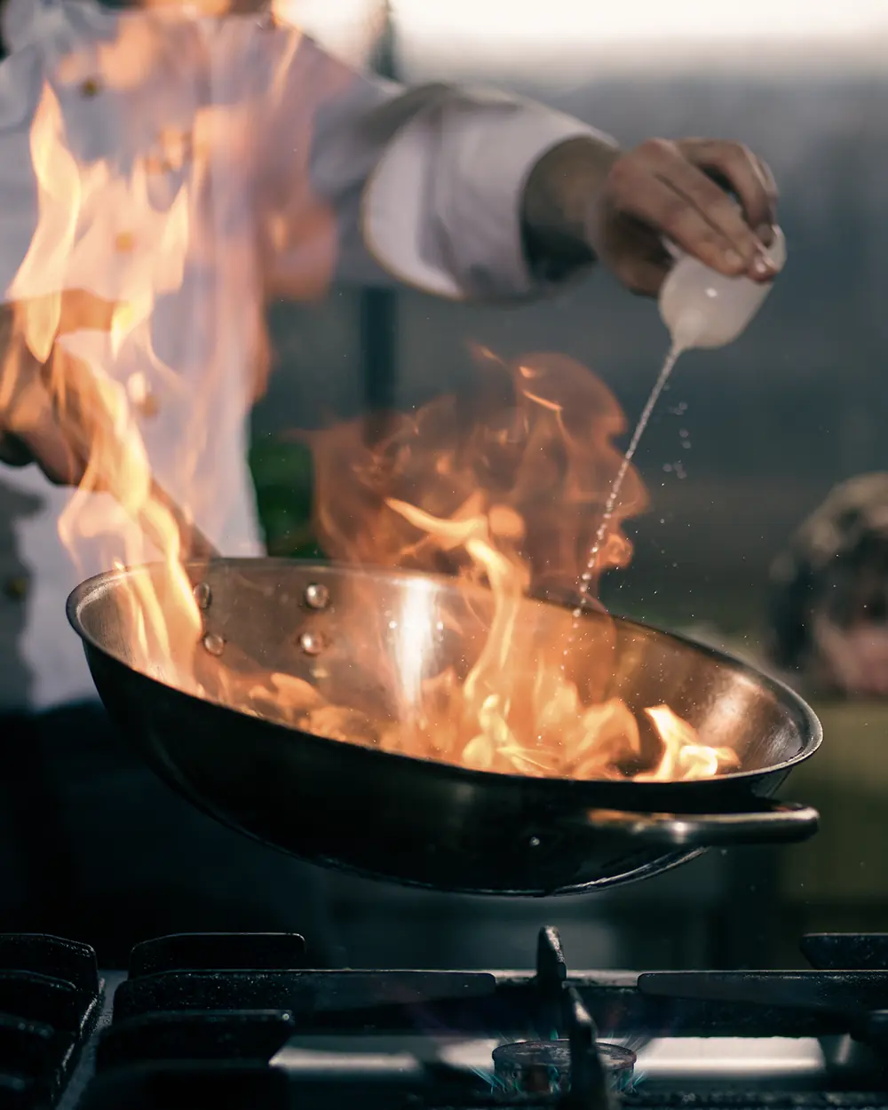 A chef finishing a dish over open flames in a pan at the EMBER pass.