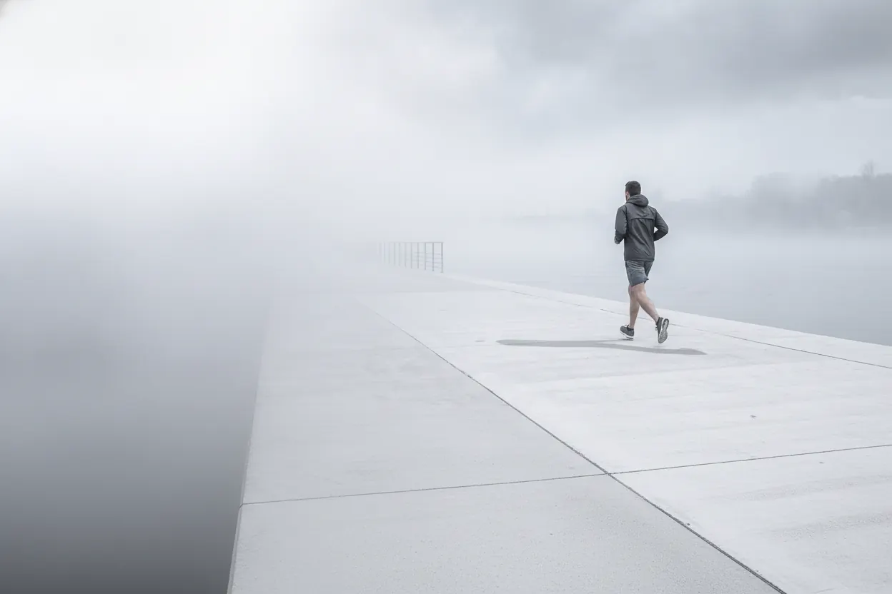 Seen from above, the runner crosses a wide pale concrete bridge alone in silver fog.