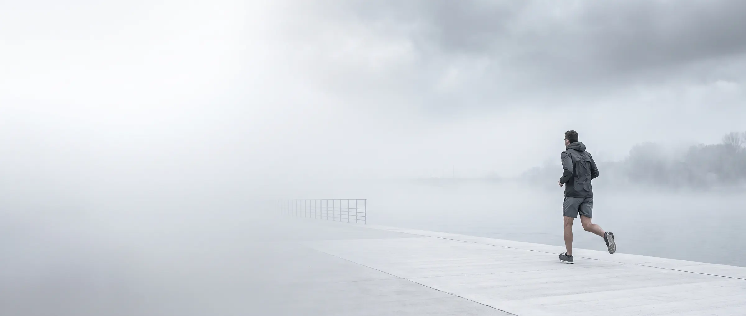 A lone runner in charcoal and grey mid-stride on a pale concrete embankment, deep in silver morning fog.