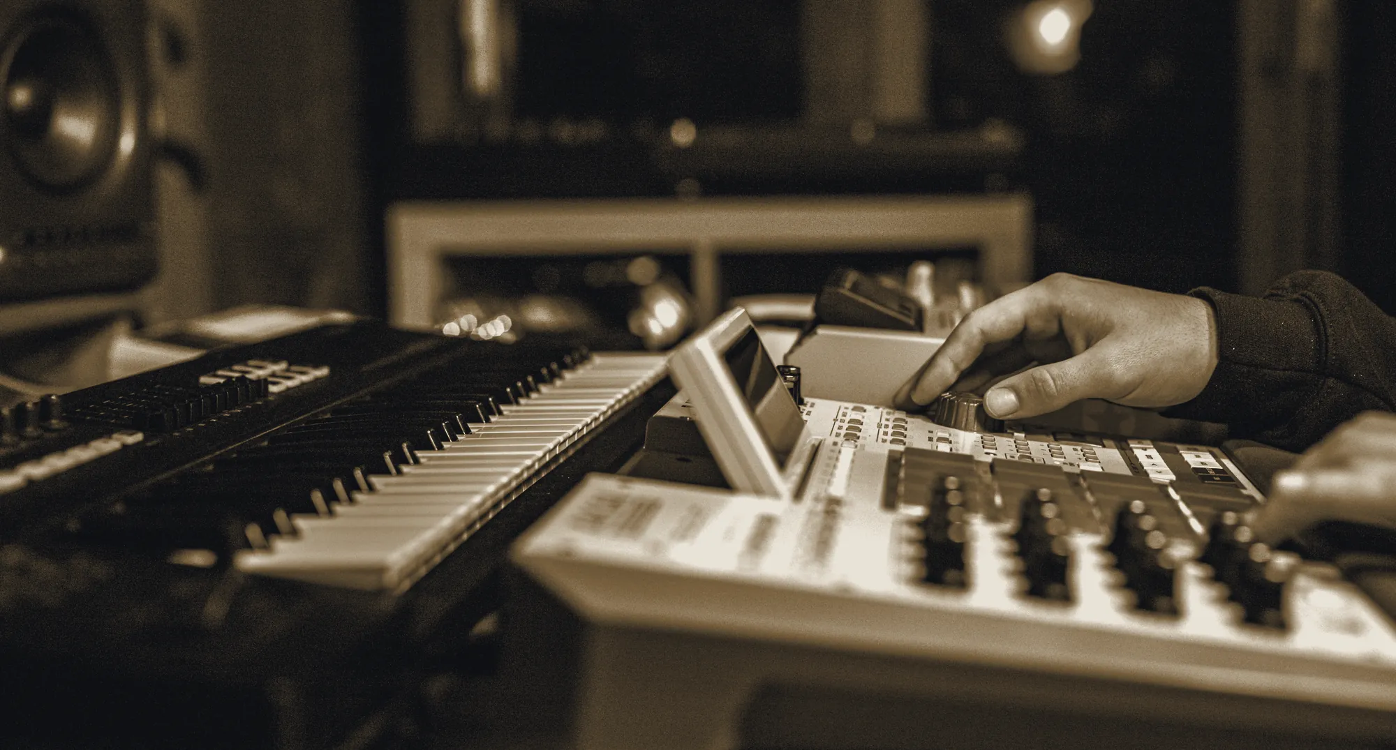 A player's hands moving across a keyboard and a drum machine in a low-lit studio.